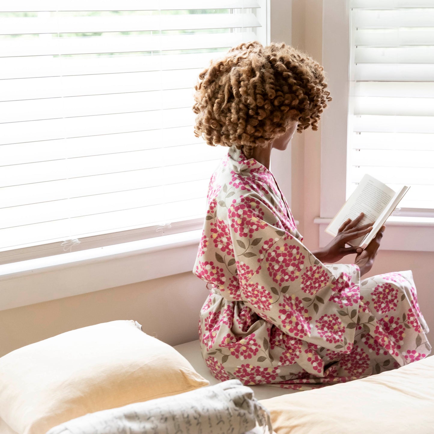 A woman with curly hair sits on a bed by a window, reading a book while wearing a pink and beige floral kimono robe.