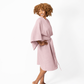 A woman with curly brown hair models a dusty pink cotton bathrobe with wide sleeves and a matching waist tie, standing against a plain white studio background.