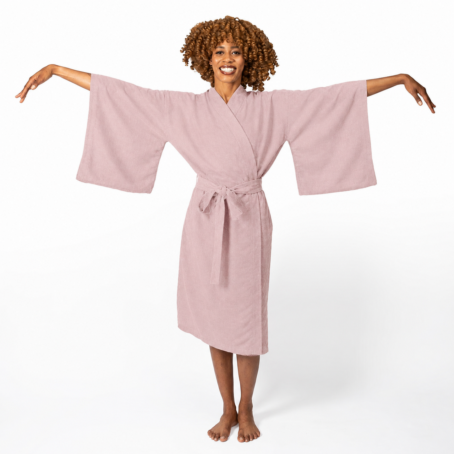 A woman with curly brown hair wearing a dusty pink cotton robe with wide sleeves and a tie belt, posing with her arms outstretched against a white studio background.