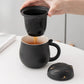 Person pouring tea from a black cat-shaped infuser into a black mug on a white surface.