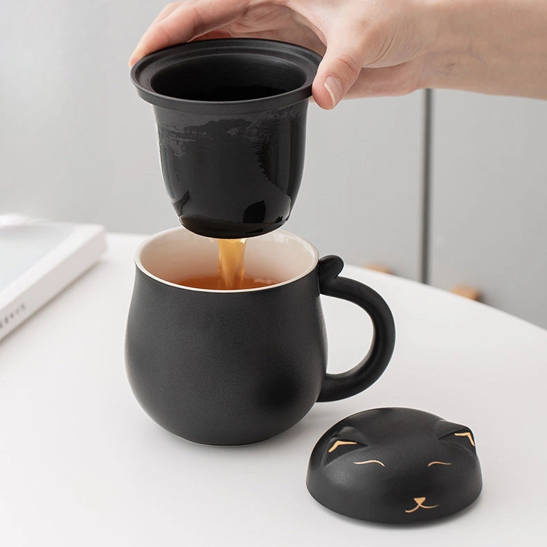 Person pouring tea from a black cat-shaped infuser into a black mug on a white surface.