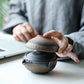 Person holding a ceramic teapot on a desk with a laptop in the background
