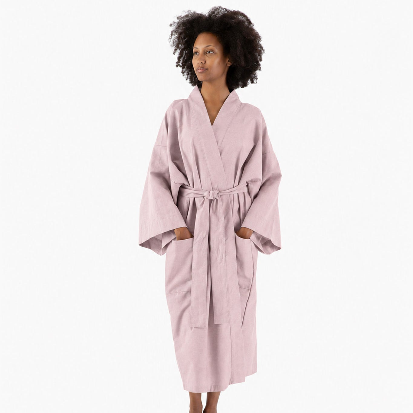 A woman wearing a light pink bathrobe with a tie belt, standing against a plain white studio background.