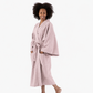A woman with dark curly hair wearing a dusty pink cotton bathrobe, standing barefoot against a solid white studio background.