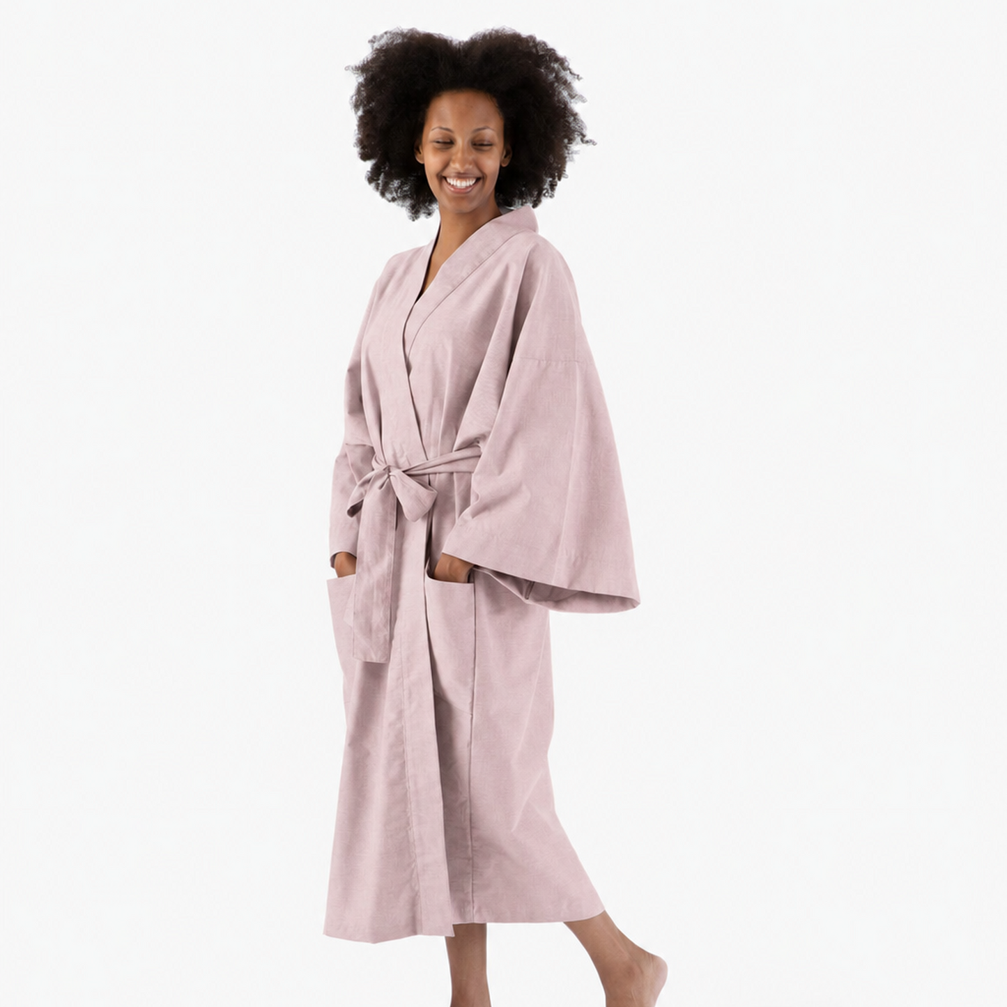 A woman with dark curly hair wearing a dusty pink cotton bathrobe, standing barefoot against a solid white studio background.
