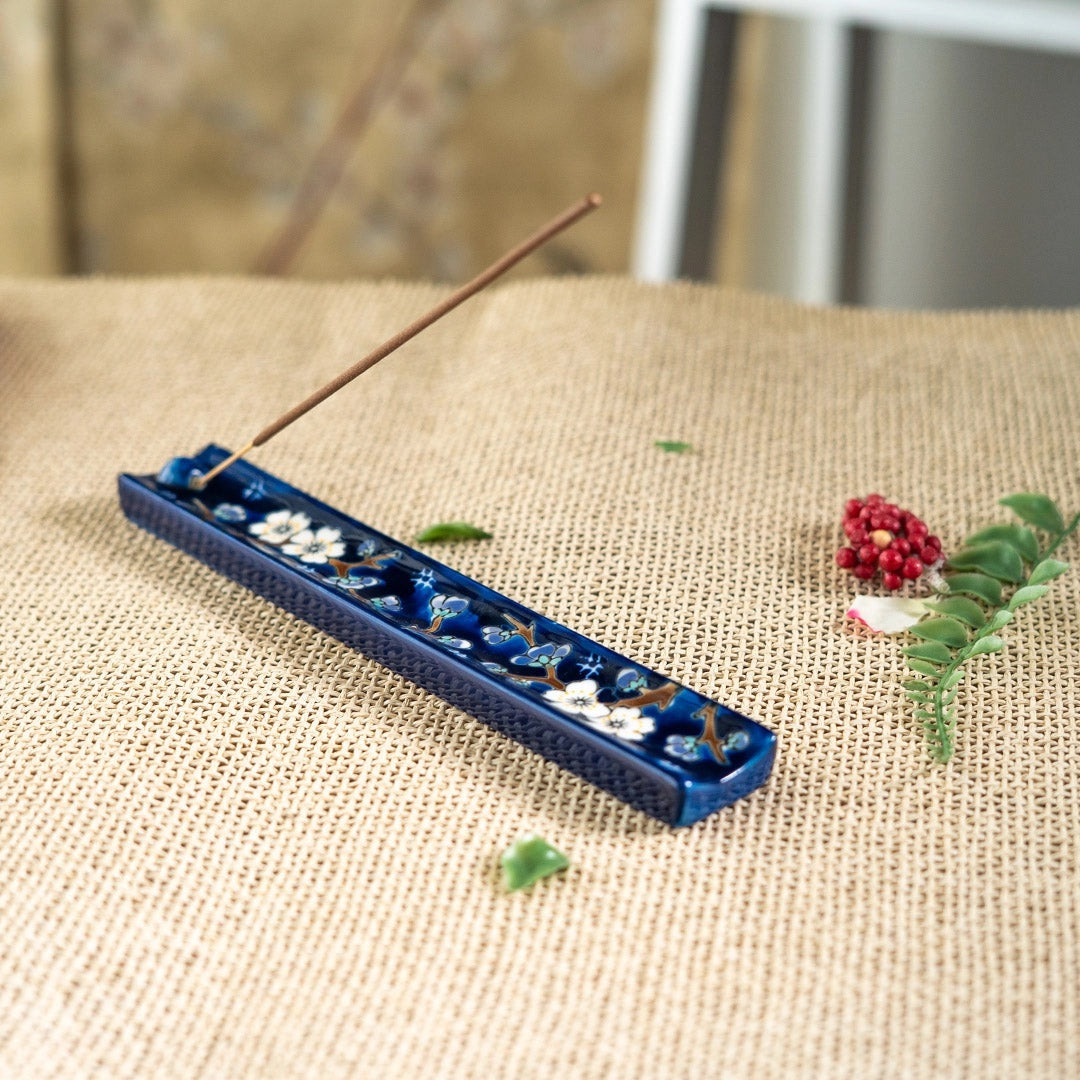 A blue ceramic incense holder with white floral details and a single incense stick inserted, placed on a beige burlap surface with small green leaves and a red flower prop nearby.
