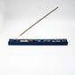 A blue rectangular incense holder with a single incense stick placed diagonally across it, photographed on a plain white studio background.
