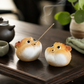 Two small pufferfish-shaped incense holders on a dark wooden tray, with smoke rising from one holding a thin incense stick. The scene includes a teapot, cup, and green plant in a softly blurred indoor setting.