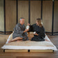 A man and a woman sitting on a low mattress on a wooden floor, both wearing matching black and white patterned robes. The man holds a white teapot while the woman holds a small white cup.