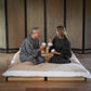 A man and a woman sitting on a low woven mat with a cream futon-style cushion, each holding a white mug in a Japanese-style room with bamboo screens and wood flooring.