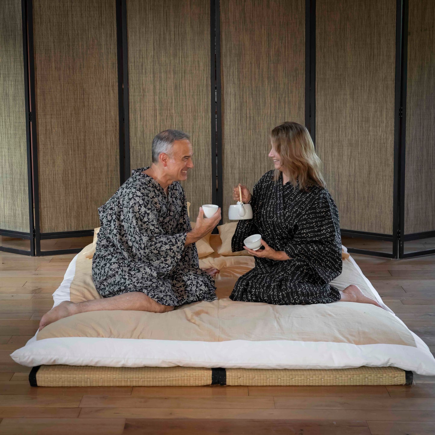 A man and a woman sitting on a low woven mat with a cream futon-style cushion, each holding a white mug in a Japanese-style room with bamboo screens and wood flooring.
