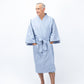A man wearing a light blue kimono-style bathrobe with wide sleeves and a tie belt, standing against a plain white studio background.