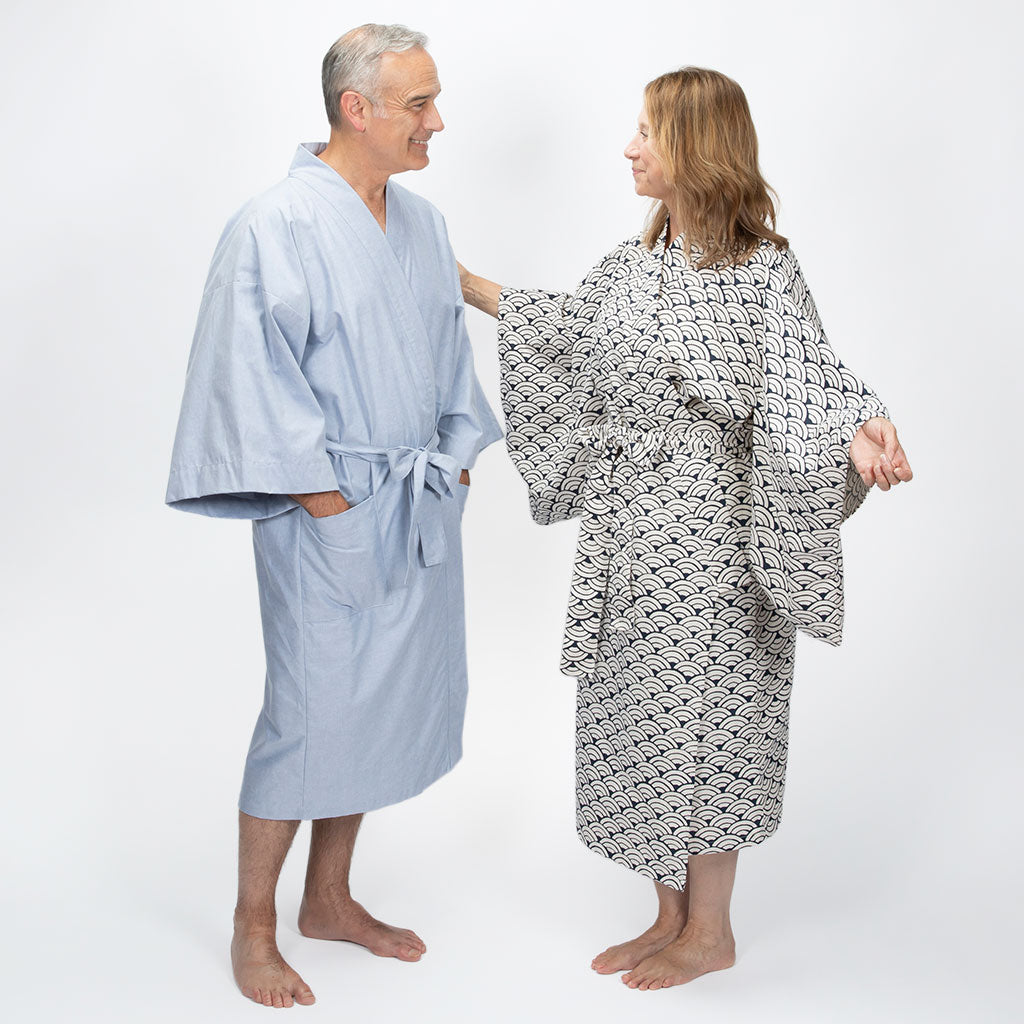 An older man and a woman standing barefoot against a white studio background, each wearing a robe. The man's robe is light blue, and the woman's robe is white with a black repeating geometric pattern.
