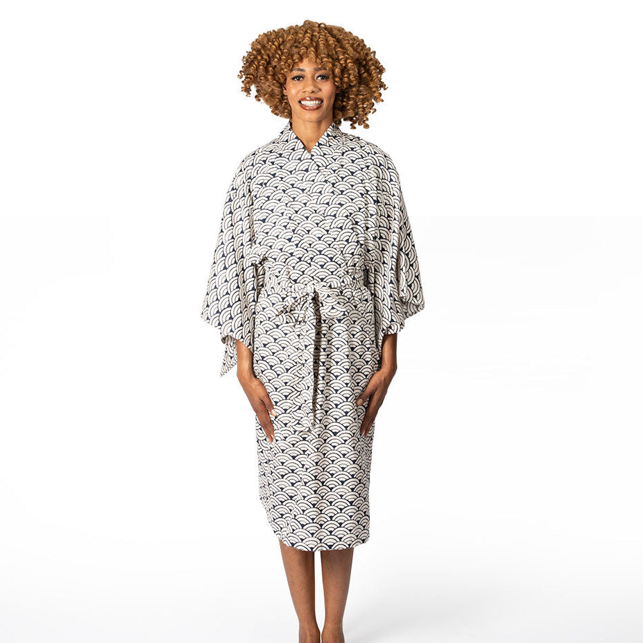 A woman with curly light brown hair stands barefoot against a plain white studio background, wearing a kimono-style robe with a black geometric pattern on a white base.