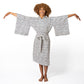 A woman with curly brown hair wearing a white and black patterned kimono robe, standing barefoot against a white studio background with her arms outstretched.