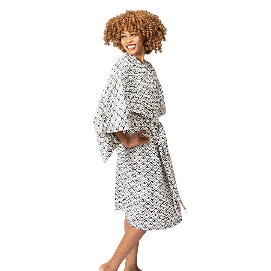 A woman modeling a white robe with a black geometric pattern, standing barefoot against a plain white studio background.