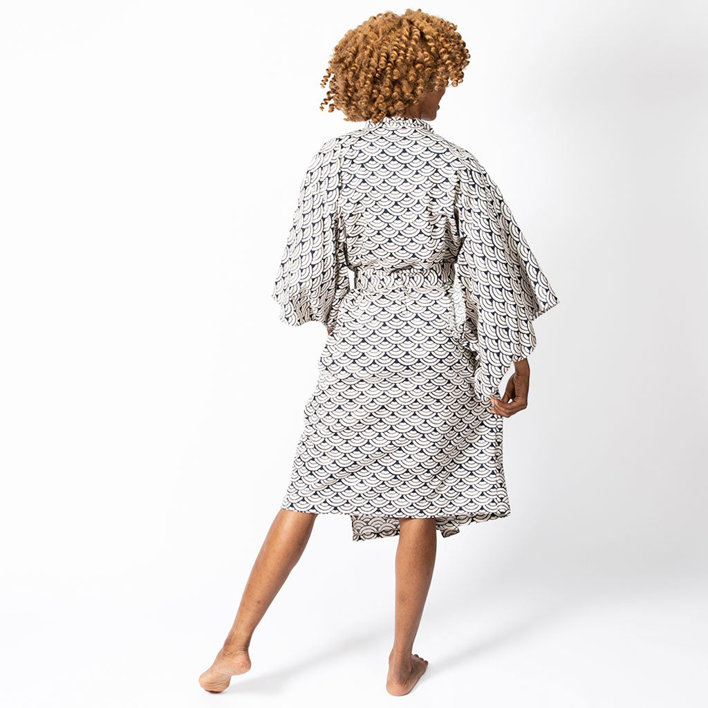 A woman shown from behind wearing a white kimono-style robe with a black geometric pattern, standing barefoot against a plain white studio background.