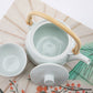 White ceramic teapot with a wooden handle and matching cup on a decorative tablecloth.