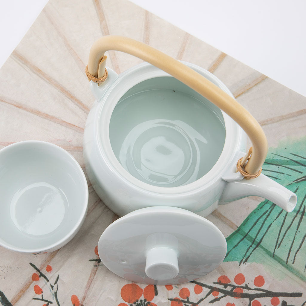 White ceramic teapot with a wooden handle and matching cup on a decorative tablecloth.