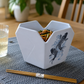 A white hexagonal ceramic bowl with a carved top opening, decorated with a blue koi fish illustration, filled with food and placed on a gray woven placemat with wooden chopsticks in front, on a wooden table.