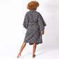 A woman with curly light brown hair is shown from behind wearing a dark navy blue and beige floral print kimono robe, standing barefoot against a plain white studio background.