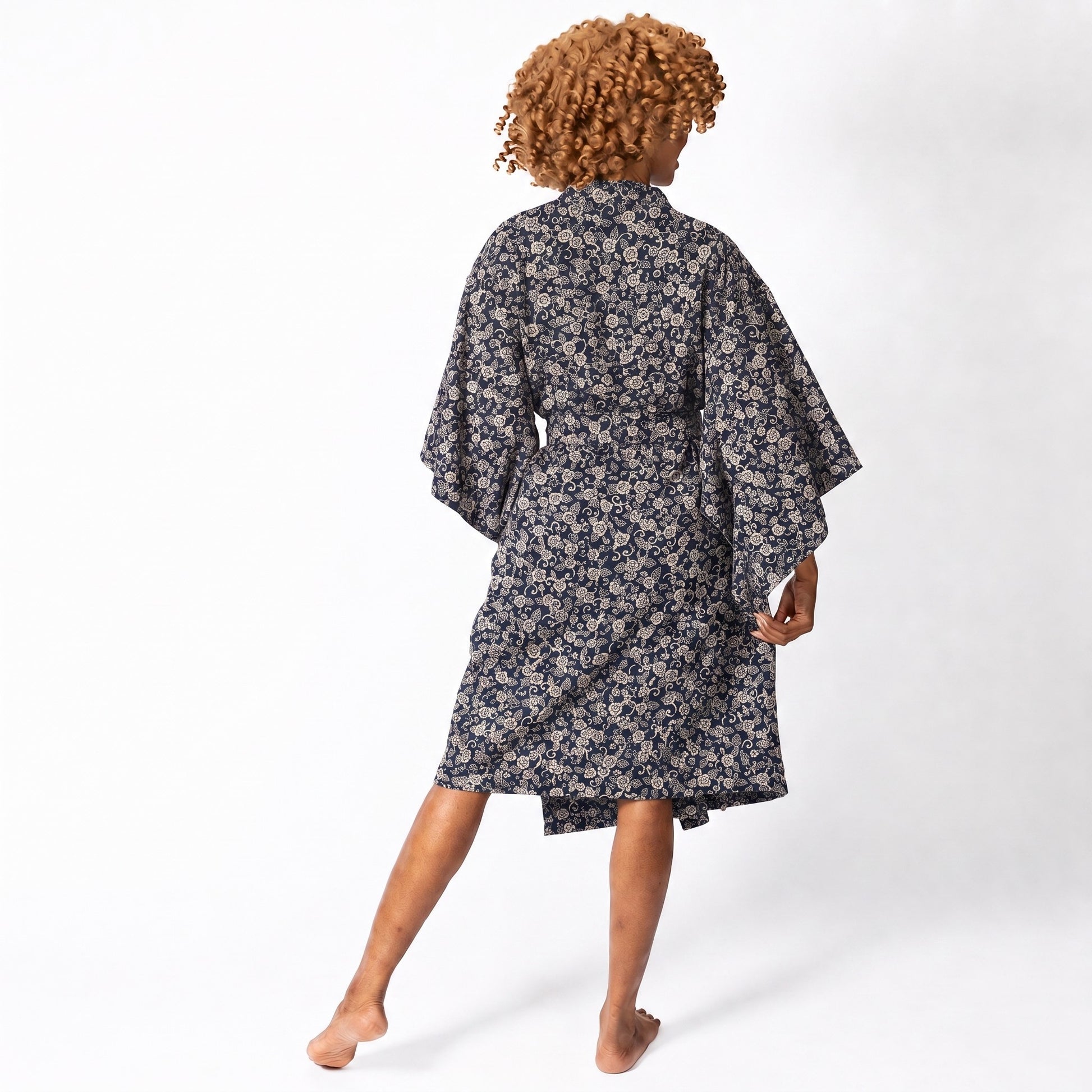 A woman with curly light brown hair is shown from behind wearing a dark navy blue and beige floral print kimono robe, standing barefoot against a plain white studio background.