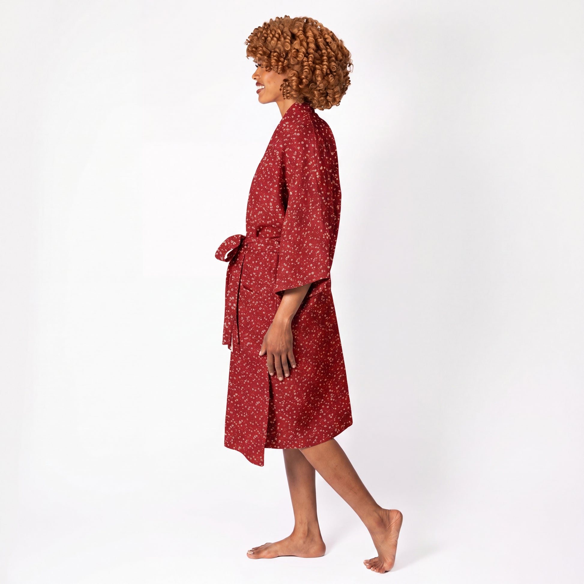 A woman with curly brown hair is shown in profile wearing a red patterned robe with a tie belt, standing barefoot against a plain white studio background.