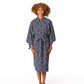 A woman with curly brown hair stands barefoot against a white studio background wearing a knee-length patterned robe with wide sleeves and a self-tie belt.