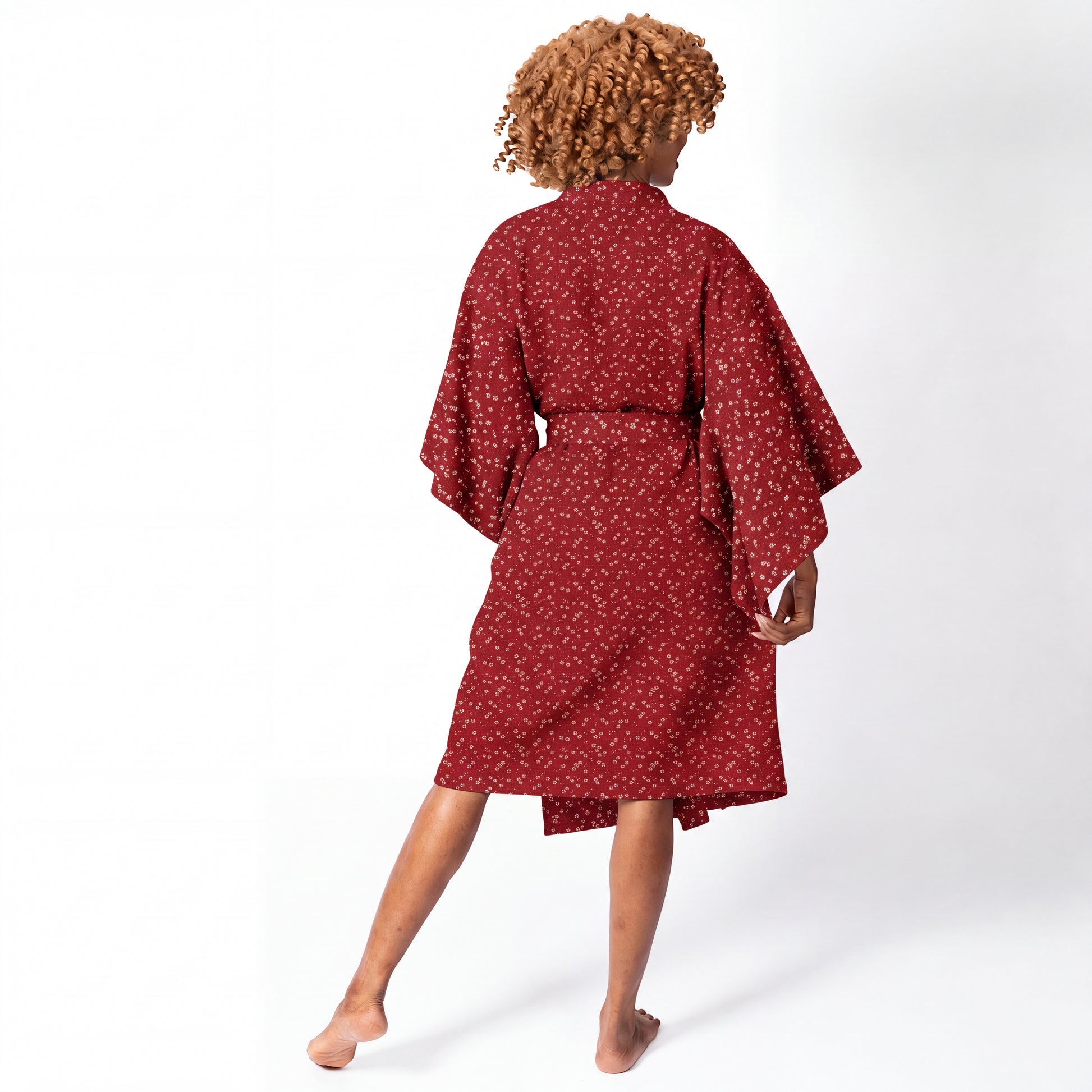 A woman shown from behind wearing a red kimono-style robe with a small white floral pattern, standing barefoot against a plain white studio background.