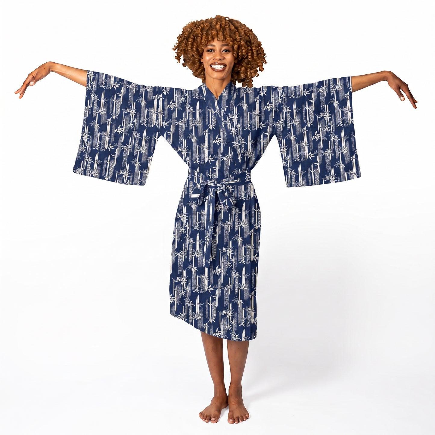 A smiling woman with curly brown hair stands barefoot against a white studio background, wearing a navy blue kimono-style robe with a white abstract pattern and wide sleeves.