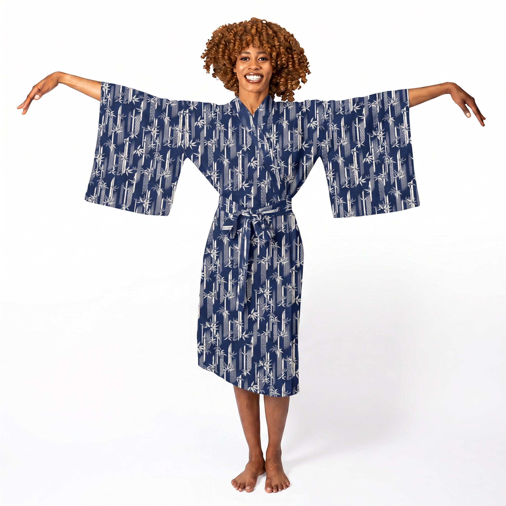 A smiling woman with curly brown hair stands barefoot against a white studio background, wearing a navy blue kimono-style robe with a white abstract pattern and wide sleeves.