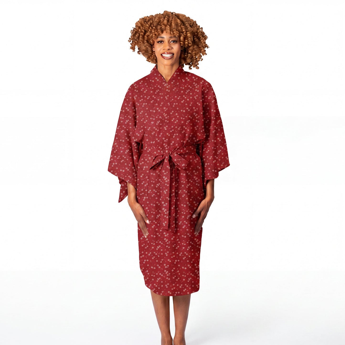 A woman with curly brown hair stands barefoot against a white studio background wearing a red kimono-style robe with a small white floral pattern and a tie belt at the waist.