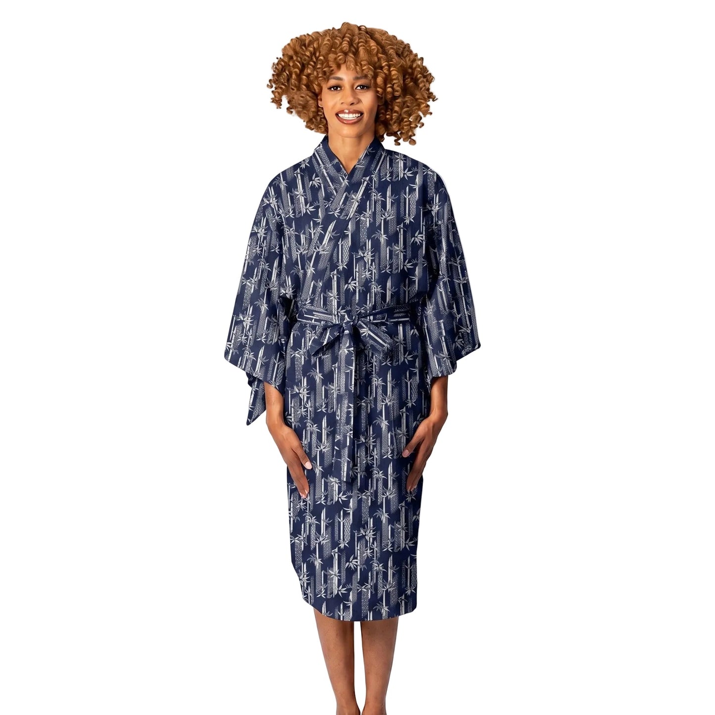 A woman with curly light brown hair stands barefoot against a white studio background wearing a navy blue kimono-style robe with a white abstract pattern and a matching tie belt.