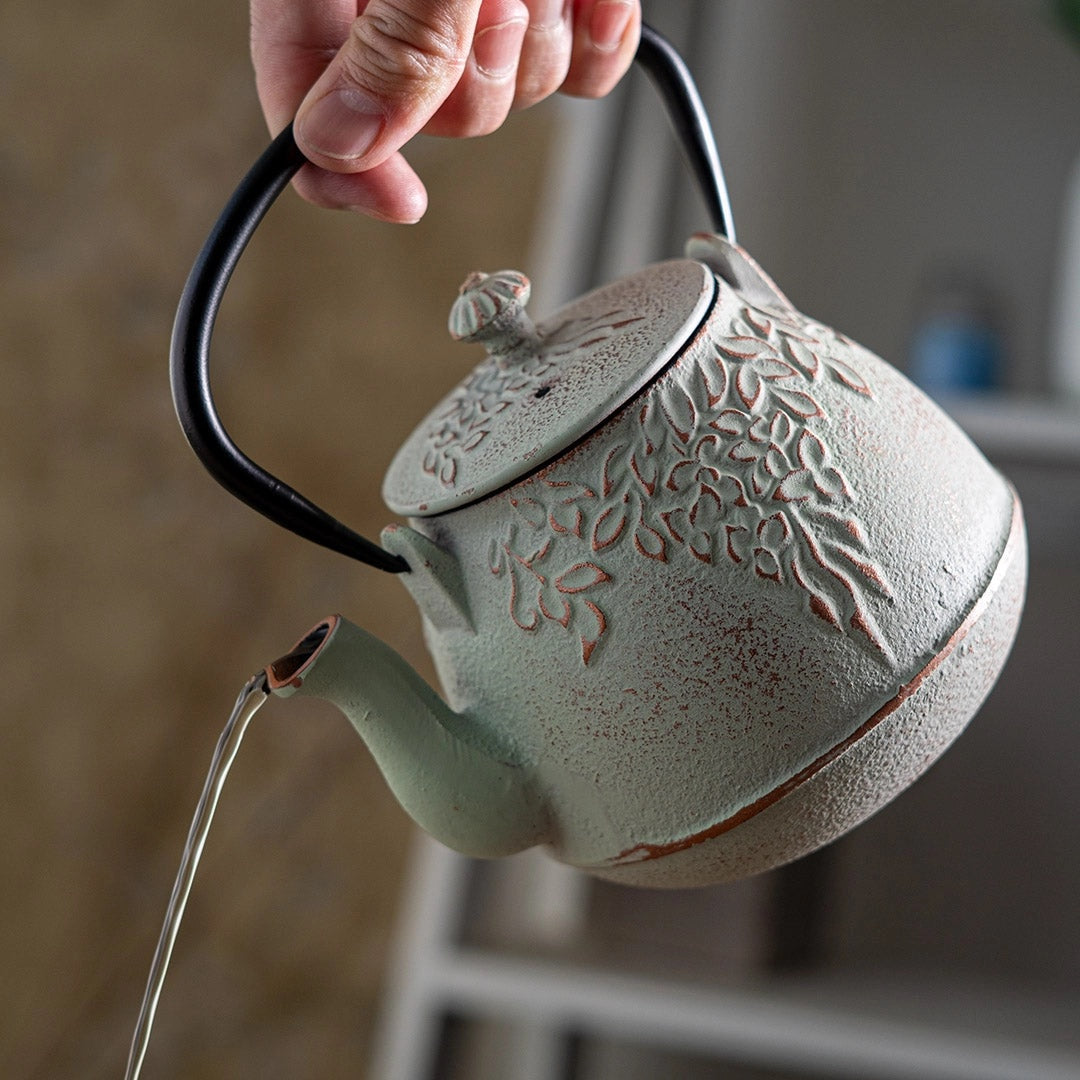A hand holds a light green cast iron teapot with a black handle and lid knob, featuring embossed floral patterns. A clear glass pouring stream is visible as water is poured from the spout, against a softly blurred indoor background.