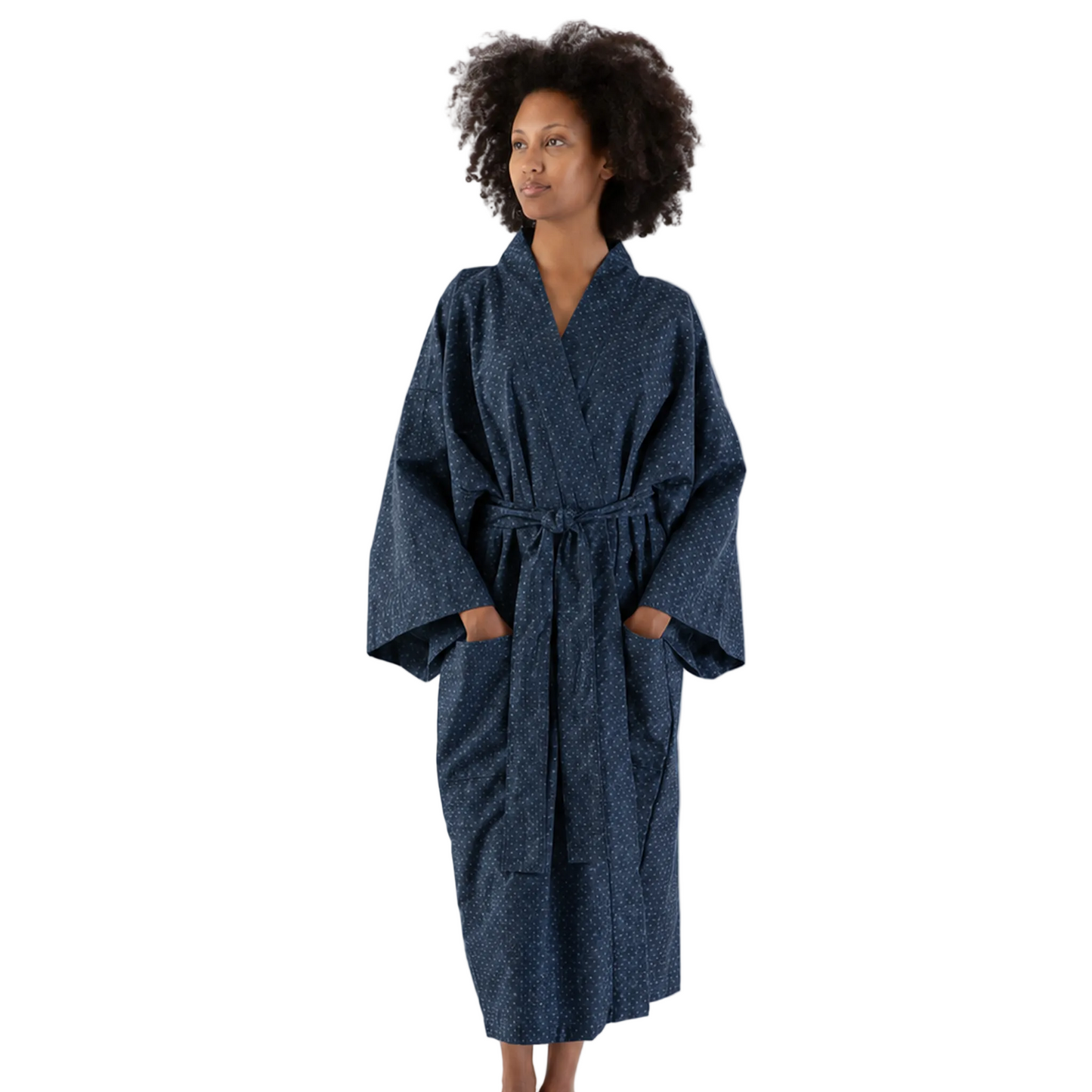 A woman stands barefoot against a plain white studio background wearing a dark navy blue patterned bathrobe with a tie belt and wide sleeves.