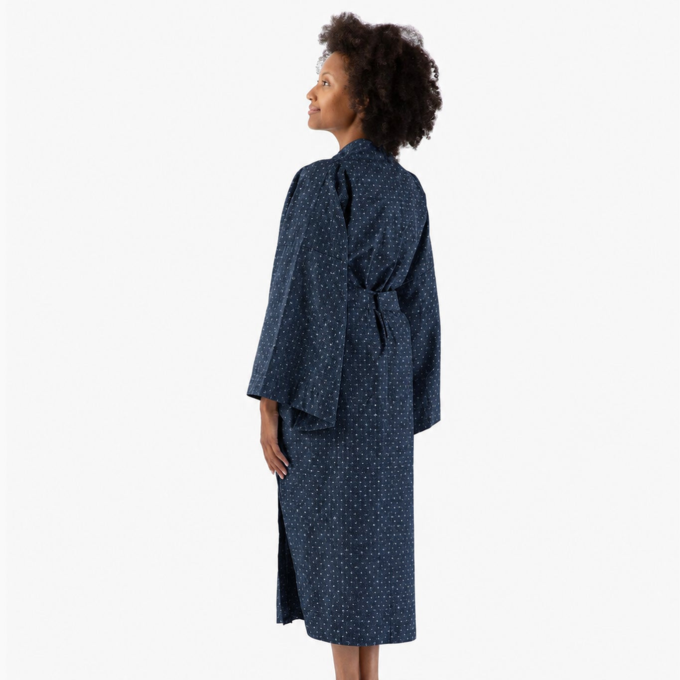 A woman stands barefoot in profile wearing a long dark navy robe with a small white allover pattern, photographed against a clean white studio background.