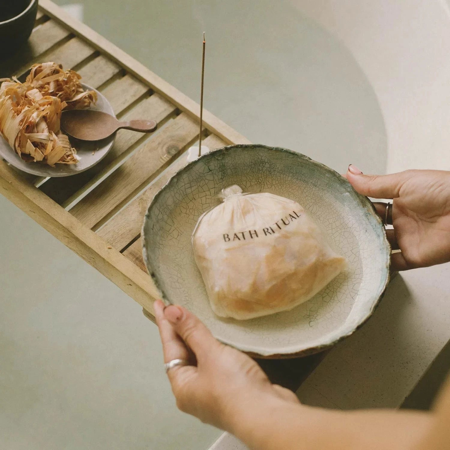 Person holding a ceramic bowl with a bag labeled 'Bath Ritual' on a wooden tray.