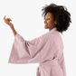 A woman with dark curly hair wearing a dusty pink kimono-style robe, shown from a side-back angle against a solid white background.