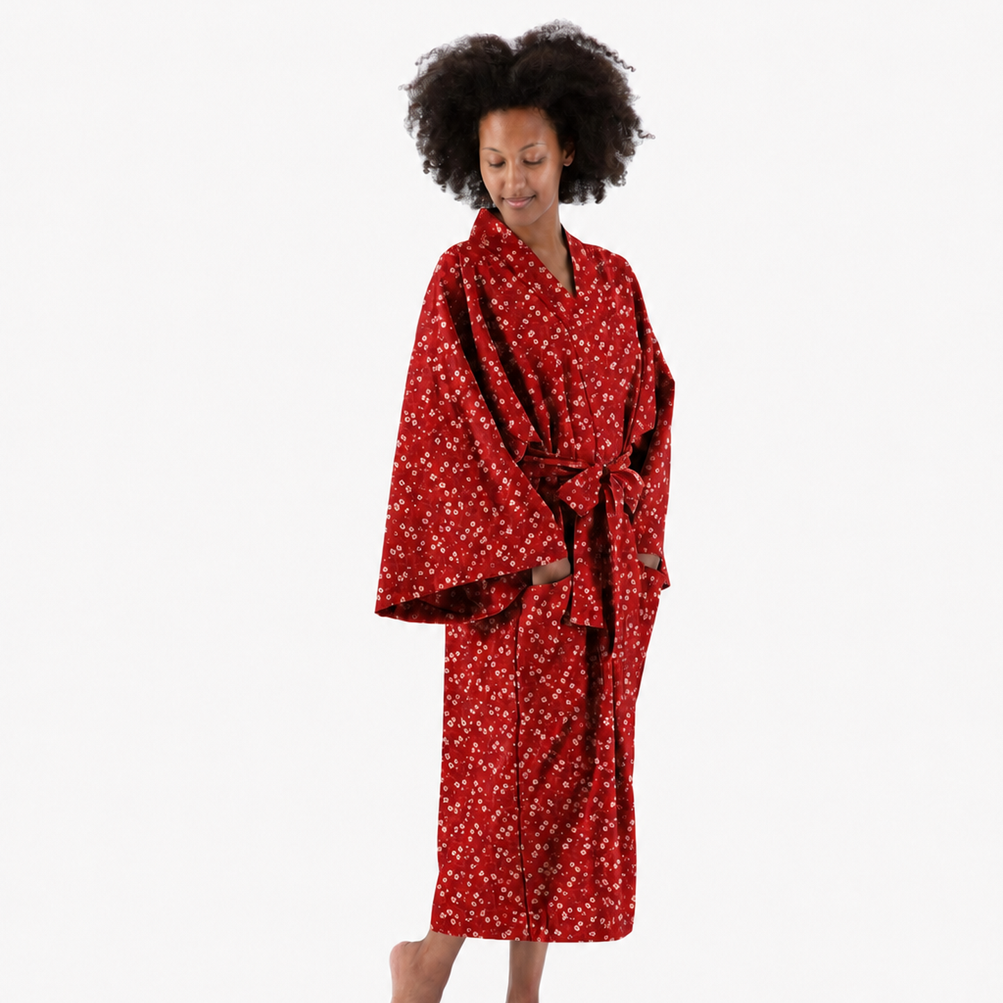 A woman with curly black hair stands barefoot against a plain white studio background, wearing a red kimono-style robe with a small white floral or ditsy print.